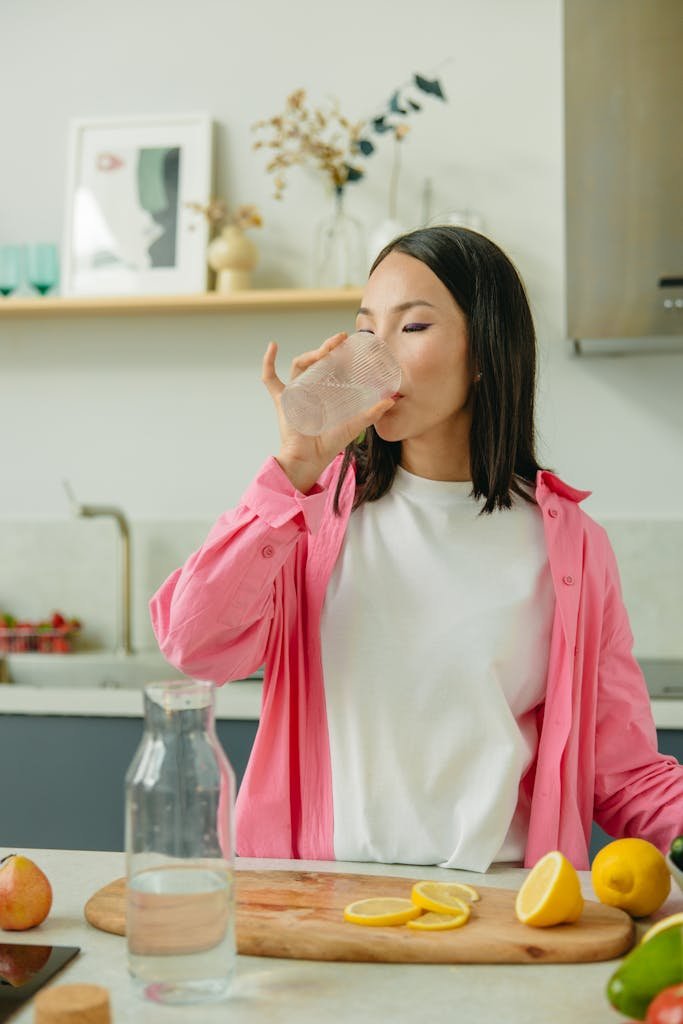 Asian woman in a pink overshirt drinks water in a kitchen setting promoting healthy lifestyle.